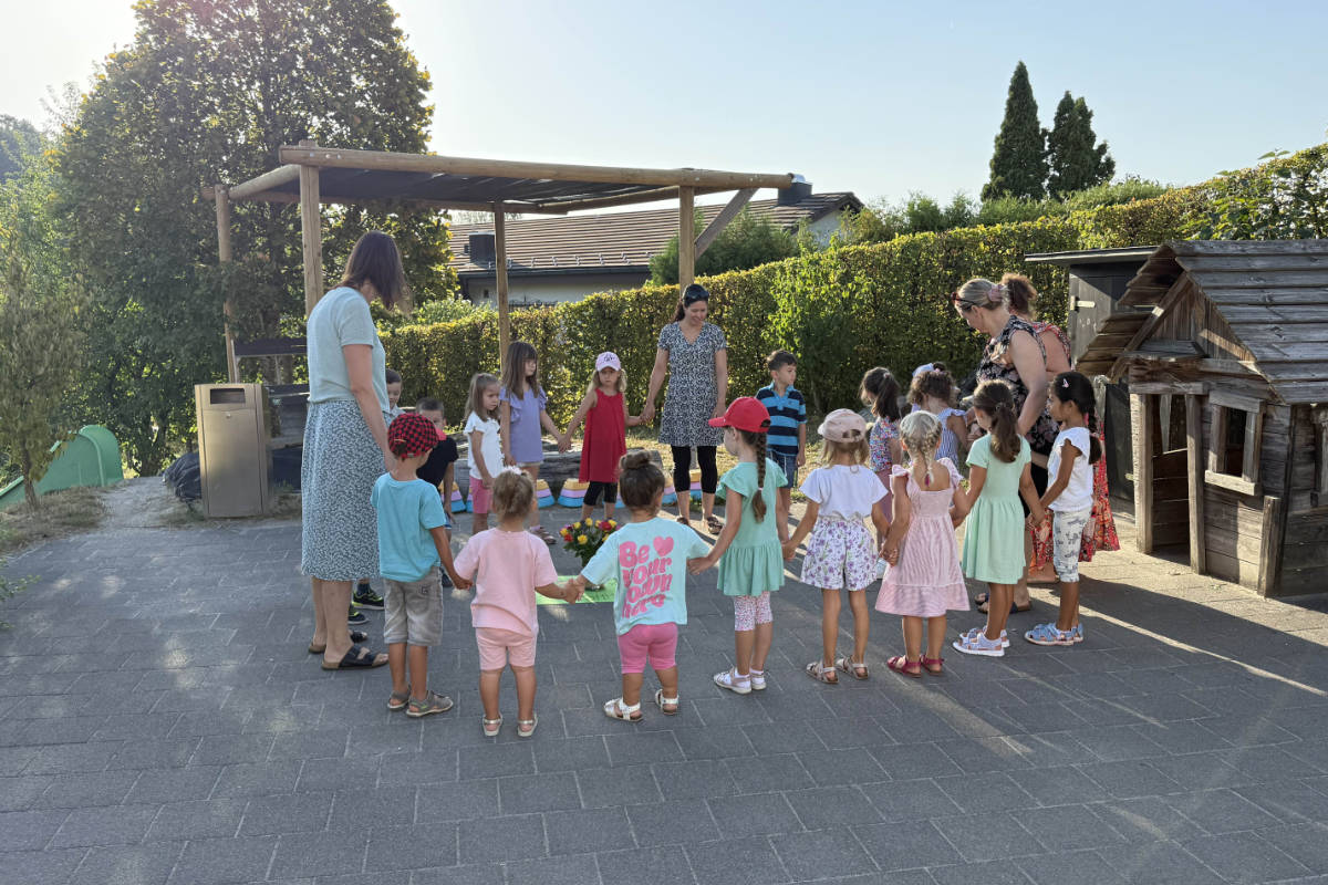 Kindergartenkinder stehen mit ihren Lehrpersonen im Kreis auf dem Pausenplatz