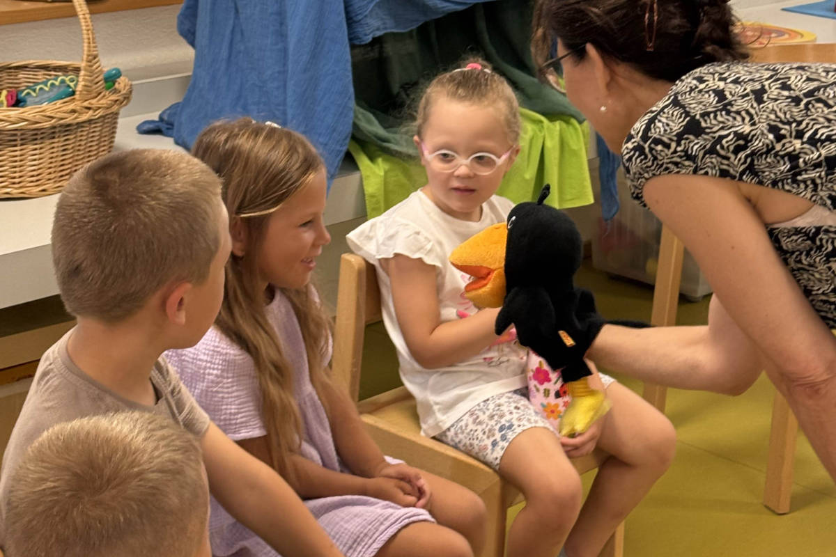 Kindergartenkinder auf Stühlen im Kreis, eine Lehrperson spielt mit Handpuppen