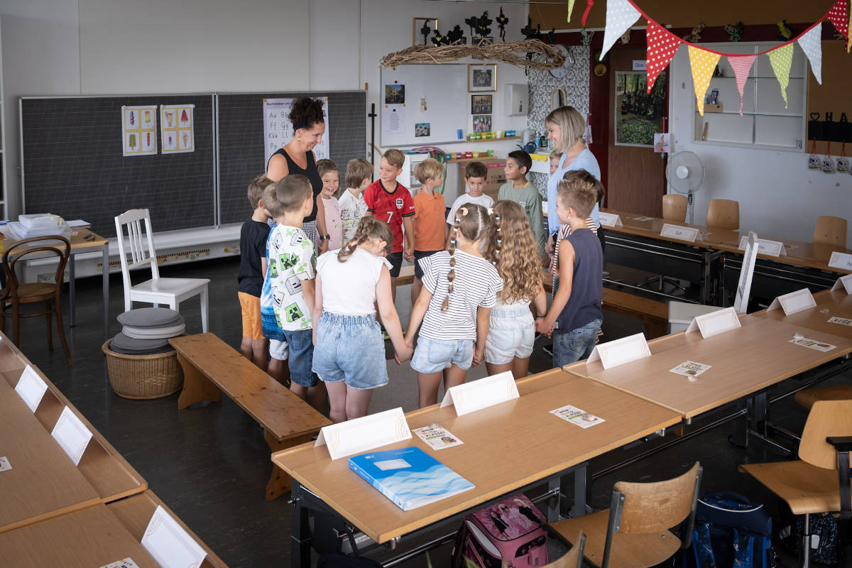 Schulkinder stehen mit ihren Lehrpersonen im Kreis im Klassenzimmer