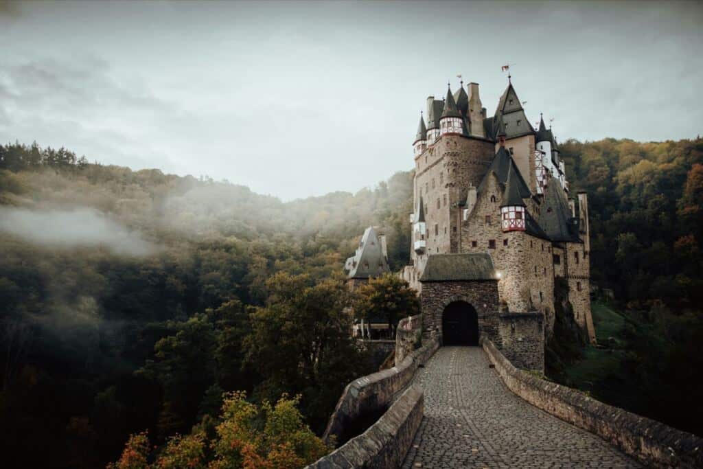 Burg mit spitzen Türmen über einem nebligen Herbstwald, davor eine gepflasterte Brücke